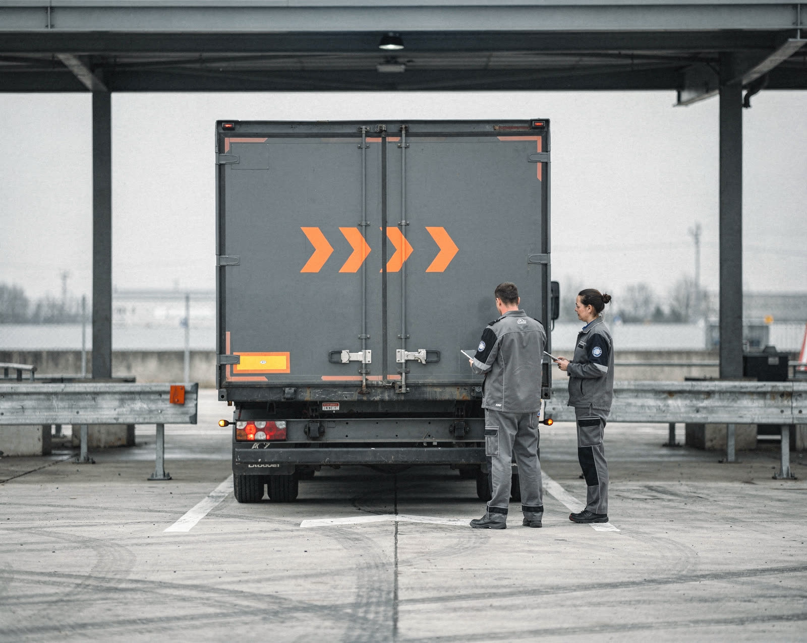 A customs officer and driver inspect the rear doors of a trailer under a checkpoint canopy